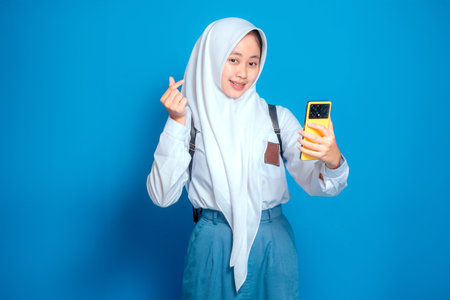 Cheerful Muslim teenage girl in school uniform smiling brightly while taking a selfie and showing finger heart gesture. Positive expression, back to school and student lifestyle.の写真素材