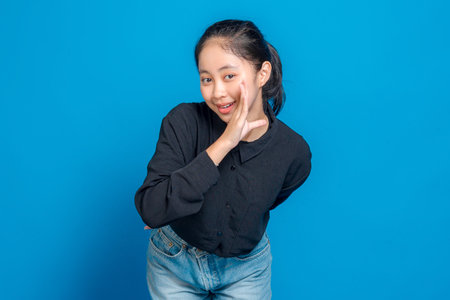 Happy young Asian woman smiling and playfully holding her hand near her mouth as if whispering or sharing a secret, expressing excitement and joy on a bright blue background.の写真素材