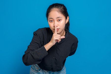Young woman with serious expression making quiet gesture by placing finger on lips, symbolizing...