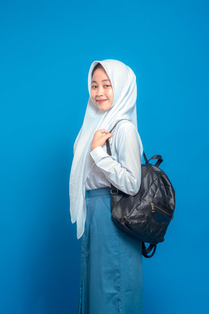 Cheerful Asian teenage girl wearing school uniform and hijab, smiling confidently while holding backpack straps. Positive and friendly facial expression on blue background.の写真素材