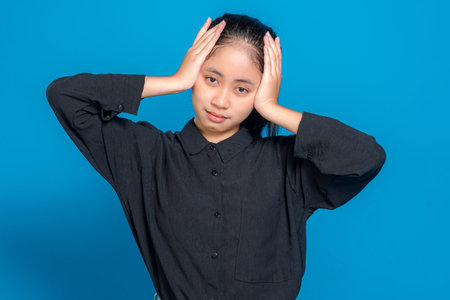 Young woman closing eyes and touching forehead with hand, showing tired or frustrated facial expression, standing against blue background, illustrating stress or mental fatigue.の写真素材