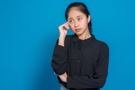 Thoughtful young Asian girl with a pensive expression, touching her temple as if thinking deeply....