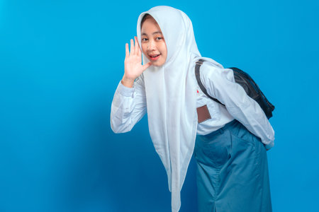 Cheerful Muslim schoolgirl in uniform and hijab making a listening gesture with playful smile and expressive face. Back to school theme on isolated blue background.の写真素材