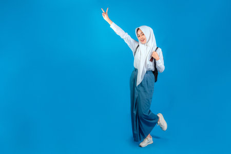 Asian Muslim teenage girl smiling cheerfully with a happy and confident expression, showing a peace sign gesture while standing on one leg, back to school education concept.の写真素材