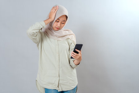 Young Asian Muslim woman wearing hijab looking at her smartphone with a frustrated facial expression and hand on head, showing stress, disappointment, or regret in casual setting.の写真素材