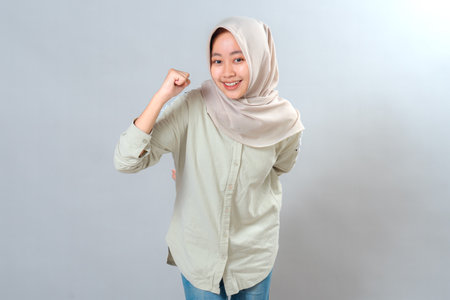 Confident young Muslim woman in hijab showing a bright smile and raising a fist in celebration. Her energetic facial expression conveys motivation, success, and determination.の写真素材