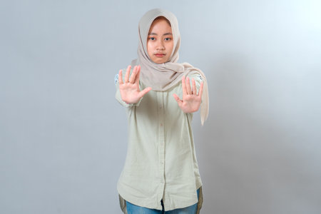 Serious young Muslim woman wearing hijab showing both hands in a stop gesture while looking directly at the camera. Her facial expression conveys refusal, warning, and concern.の写真素材