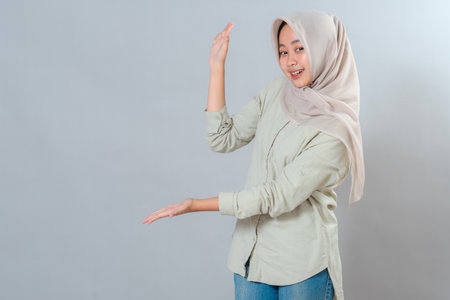Cheerful young Muslim woman in hijab smiling and gesturing with both hands to present or highlight something. Her friendly and confident expression suits advertising and marketing needs.の写真素材