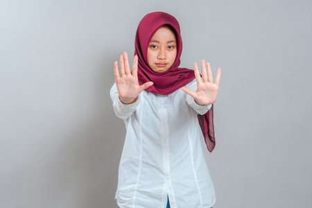 Serious young woman in maroon hijab and white shirt showing stop gesture with her hand. Her face expresses firmness and warning, ideal for campaigns about boundaries or awareness.の写真素材