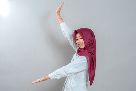 Cheerful young woman in maroon hijab and white shirt smiling while making a welcoming hand gesture. Her face radiates friendliness and charm, perfect for positive advertising content.の写真素材