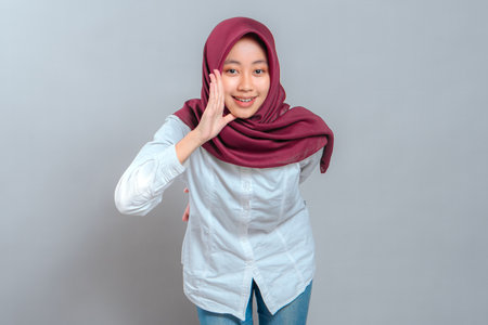 Young woman in maroon hijab smiling brightly while cupping her hand near her mouth, showing an expressive and friendly face as if calling or sharing a fun secret with enthusiasm.の写真素材