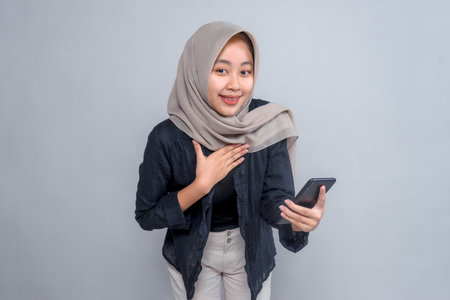 Smiling Asian woman in hijab looking at smartphone, hand on chest in grateful or touched gesture,...