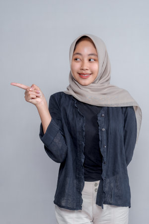 Portrait of a cheerful young Asian Muslim woman wearing hijab, smiling while pointing to the side, standing against plain background, perfect for advertisement or copy space content.の写真素材