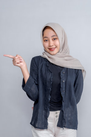 Confident young Asian Muslim woman in hijab smiling while pointing sideways with her index finger, posing against a plain background, perfect for promotional or marketing content.の写真素材