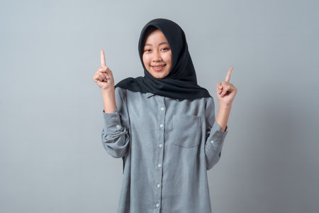 Portrait of a smiling young Asian Muslim woman in a black hijab and grey shirt, pointing both index fingers upward, suggesting copy space or important information above.の写真素材