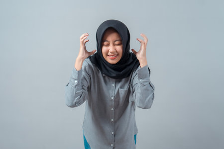 Young Asian Muslim woman wearing a black hijab and gray shirt showing a stressed, frustrated expression with clenched hands near her head, eyes closed, isolated on gray background.の写真素材