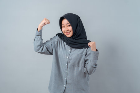 Confident young Muslim woman in hijab and grey shirt smiling and showing arm muscles in a strong pose, representing empowerment, strength, and positivity on plain background.の写真素材