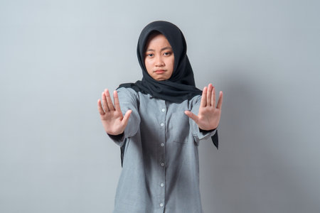 Serious young Asian Muslim woman wearing a hijab and gray shirt making a stop gesture with both hands, standing against a neutral gray background, expressing refusal.の写真素材