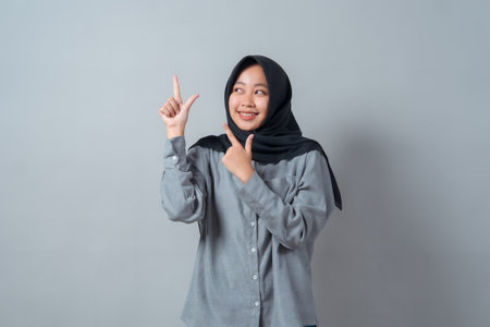 Smiling young Asian Muslim woman in hijab and gray shirt, pointing with both hands while looking up, showing a cheerful expression against a plain gray background.の写真素材