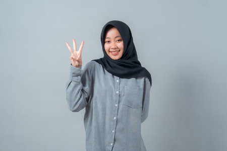 Happy young Muslim woman wearing a black hijab and gray shirt smiling while showing three fingers with her right hand, standing confidently in front of a plain background.の写真素材