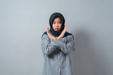 Young Asian Muslim woman wearing a hijab and gray shirt, standing against plain background, making an âXâ sign with her arms while showing a serious face to express rejection.の写真素材