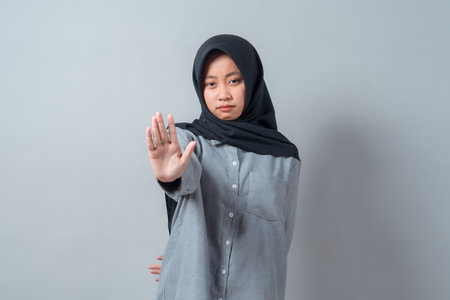 Confident young Asian Muslim woman in a hijab and gray shirt showing a stop gesture with one hand, looking serious while standing against a neutral gray background.の写真素材