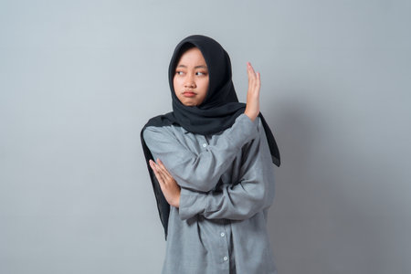 Serious young Muslim woman in hijab making a rejecting hand gesture with displeased facial expression, wearing a grey shirt, standing against a neutral light grey background.の写真素材
