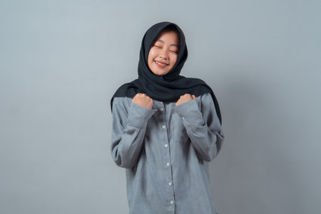 Young Muslim woman in black hijab and gray shirt smiling with eyes closed and fists clenched near...