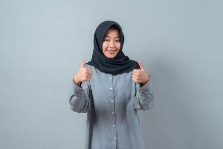 Cheerful young Asian Muslim woman in a black hijab and gray shirt smiling while giving two thumbs up gesture, representing positivity, motivation, approval, and success.の写真素材