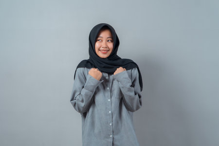 Smiling young Muslim woman in black hijab and gray shirt, clenching fists near her chest in a shy but confident gesture. Concept of optimism, hope, strength, and ambition.の写真素材