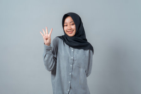 Smiling young Muslim woman in a black hijab and gray shirt standing against a plain background, holding up four fingers with her right hand in a confident pose.の写真素材