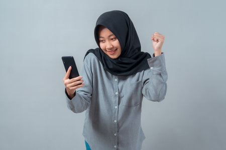 Young Muslim woman in black hijab smiling and raising fist in excitement while holding smartphone,...
