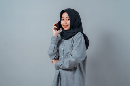 Portrait of a young Asian Muslim woman wearing a black hijab and grey shirt, smiling confidently while holding a smartphone near her ear, standing against a plain grey background.の写真素材