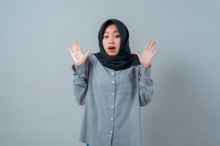 A young Asian Muslim woman wearing a hijab and grey shirt looks surprised with raised hands and open mouth, standing in front of a plain background, expressing shock.の写真素材