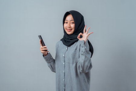 Portrait of a smiling young Asian Muslim woman in a black hijab and grey shirt holding a smartphone and making an OK hand gesture, standing confidently against a plain light background.の写真素材