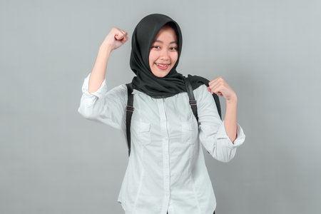 Cheerful Muslim teenage girl in a black hijab and white shirt smiles confidently while raising both fists, expressing school spirit, motivation, and academic readiness.の写真素材