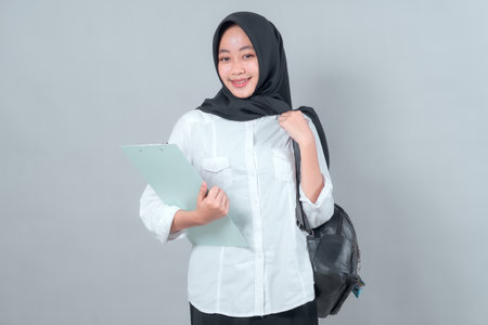 Portrait of a young Muslim woman in a black hijab and white shirt, smiling while holding a clipboard and wearing a backpack, expressing confidence in a back-to-school theme.の写真素材