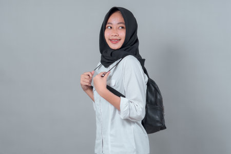 A cheerful young Asian woman in a black hijab and white shirt, holding her backpack straps and looking sideways with a smile, poses against a plain grey background.の写真素材