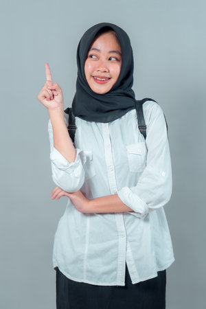 Cheerful Muslim teenage girl in black hijab and white shirt smiling while pointing upward with index finger. Confident student expressing bright idea on plain gray background.の写真素材