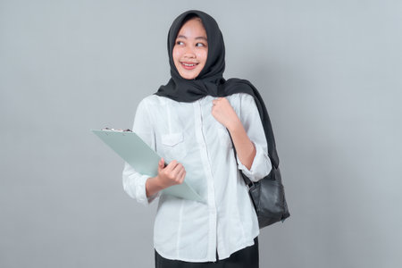 A cheerful young Muslim woman in a black hijab and white shirt, holding a clipboard and backpack, looks to the side confidently, symbolizing education and backtoschool enthusiasm.の写真素材