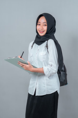 Happy Muslim teenage girl in a black hijab and school uniform smiling while writing on a clipboard. She carries a backpack, ready to face a new school day.の写真素材