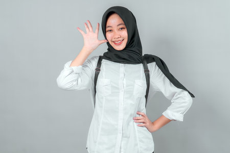 Smiling Asian Muslim teenage girl in a black hijab and white shirt, posing confidently with a backpack and showing five fingers, symbolizing excitement for back to school time.の写真素材