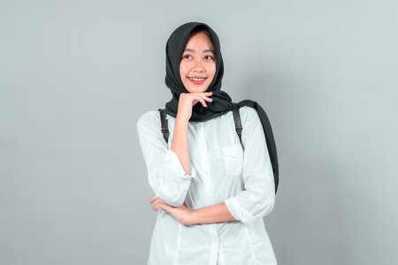 Portrait of happy Asian Muslim teenage girl in hijab with backpack, looking up with smile, symbolizing optimism, education, and back to school on plain gray background.の写真素材