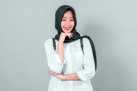 Portrait of a cheerful Muslim teenage girl wearing a black hijab and white shirt, holding her chin with one hand, posing confidently in a back to school theme on gray background.の写真素材