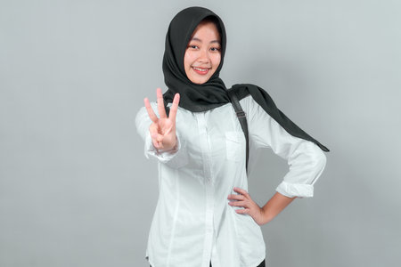 Happy Muslim schoolgirl wearing a black hijab and white shirt, smiling and showing three fingers with hand forward. Confident teenage student ready for back to school season.の写真素材