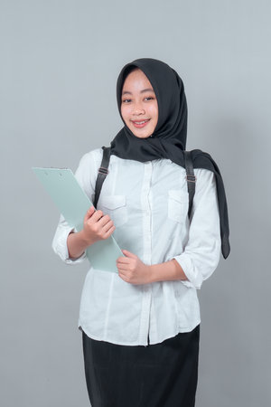 Portrait of a smiling Asian Muslim student wearing a black hijab, white shirt, and backpack, holding a clipboard confidently. Education, back to school concept.の写真素材