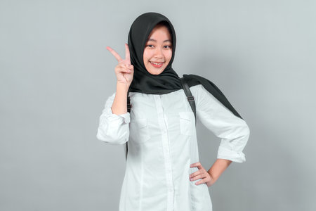 Smiling Asian Muslim schoolgirl wearing a white shirt, black hijab, and backpack, showing a peace sign with her fingers. Studio shot with gray background. Back to school concept.の写真素材