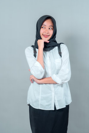 Portrait of a cheerful Muslim teenage girl in black hijab and white shirt, looking aside with a smile, standing confidently with a backpack, ready for school.の写真素材