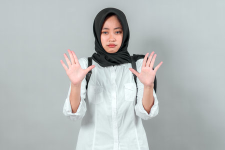 Muslim student girl wearing hijab and backpack raising hands with serious expression, isolated on gray background. Education, warning, back to school concept.の写真素材