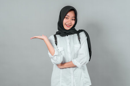 Happy Asian Muslim student in a black hijab and white shirt smiling while presenting with open hand gesture, showing confidence and excitement for back to school, on gray.の写真素材
